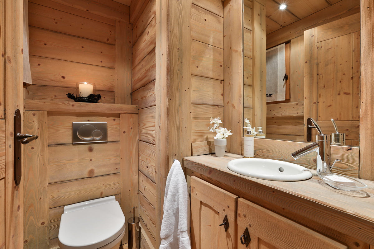 A rustic wooden bathroom interior with a white toilet, white sink basin, chrome faucet, mirror, and light wood cabinetry. A white towel hangs from the sink area, and a small shelf with a lit candle is visible on the left.