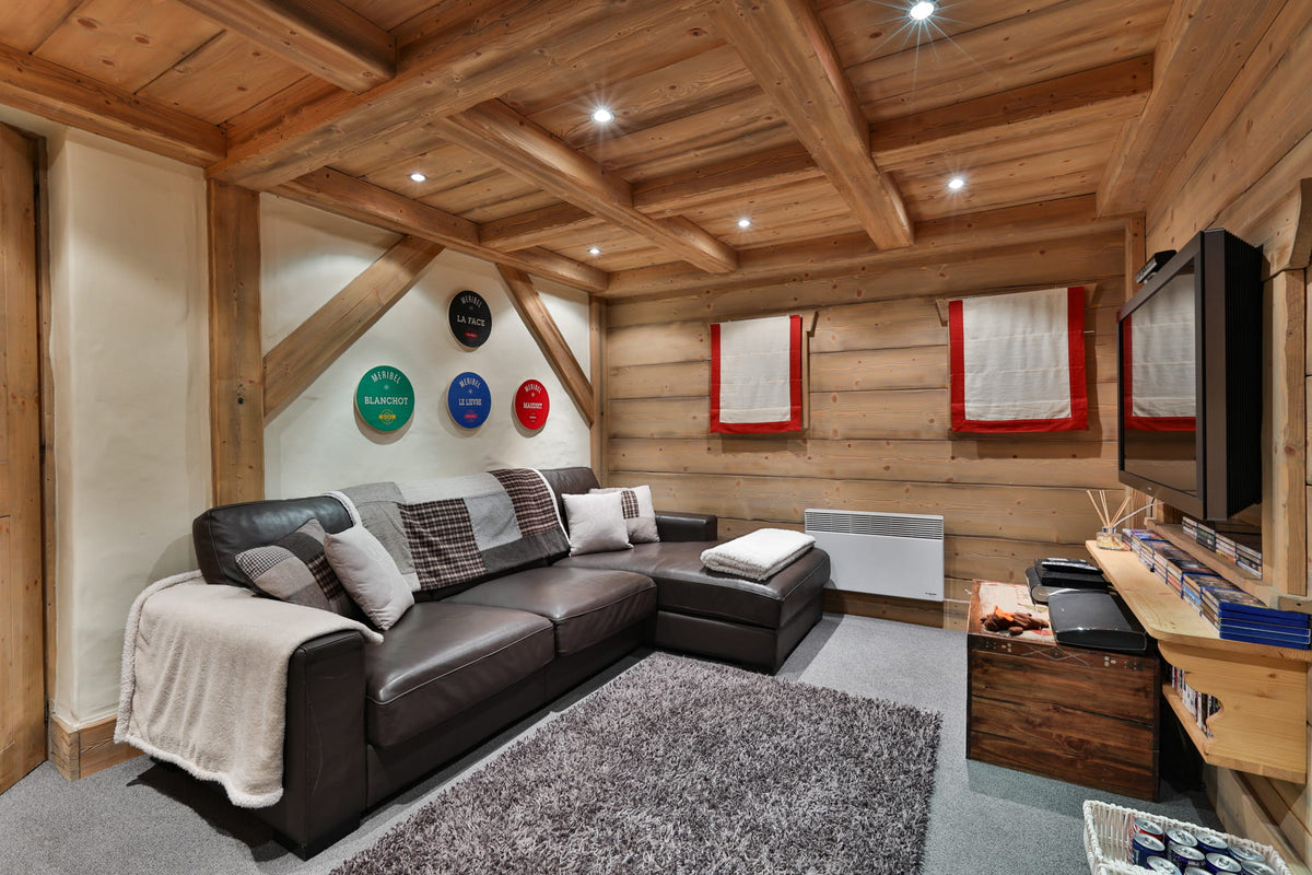 A modern ski chalet living room featuring a dark brown leather sectional sofa with throw pillows, a gray shag rug, wood-paneled walls and ceiling with exposed beams, and a TV mounted on the right wall.