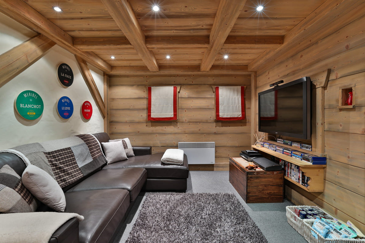 A cozy ski chalet-style living room with a dark brown leather sectional sofa, a gray shag rug, wood-paneled walls and ceiling with exposed beams, a wall-mounted TV, and decorative circular signs on the left wall.