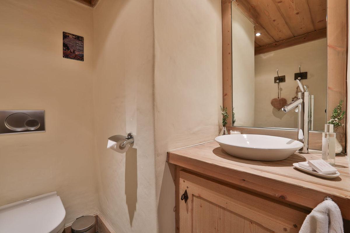 A rustic bathroom interior with a wooden vanity, white vessel sink, wall-mounted mirror, and warm beige plaster walls.