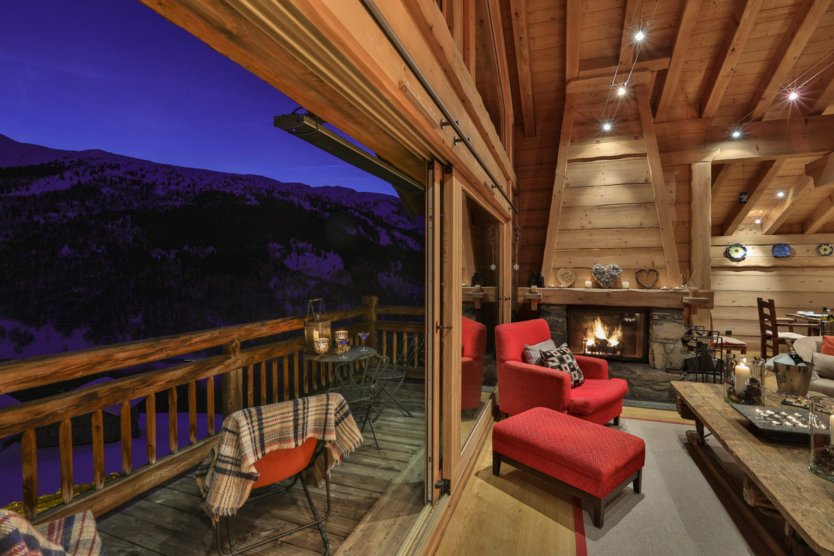 A cozy mountain chalet interior with large sliding glass doors opening onto a deck overlooking a purple-toned mountain landscape at dusk. The room features red upholstered armchairs and an ottoman, a lit fireplace, wooden ceiling beams, and a rustic wood coffee table.