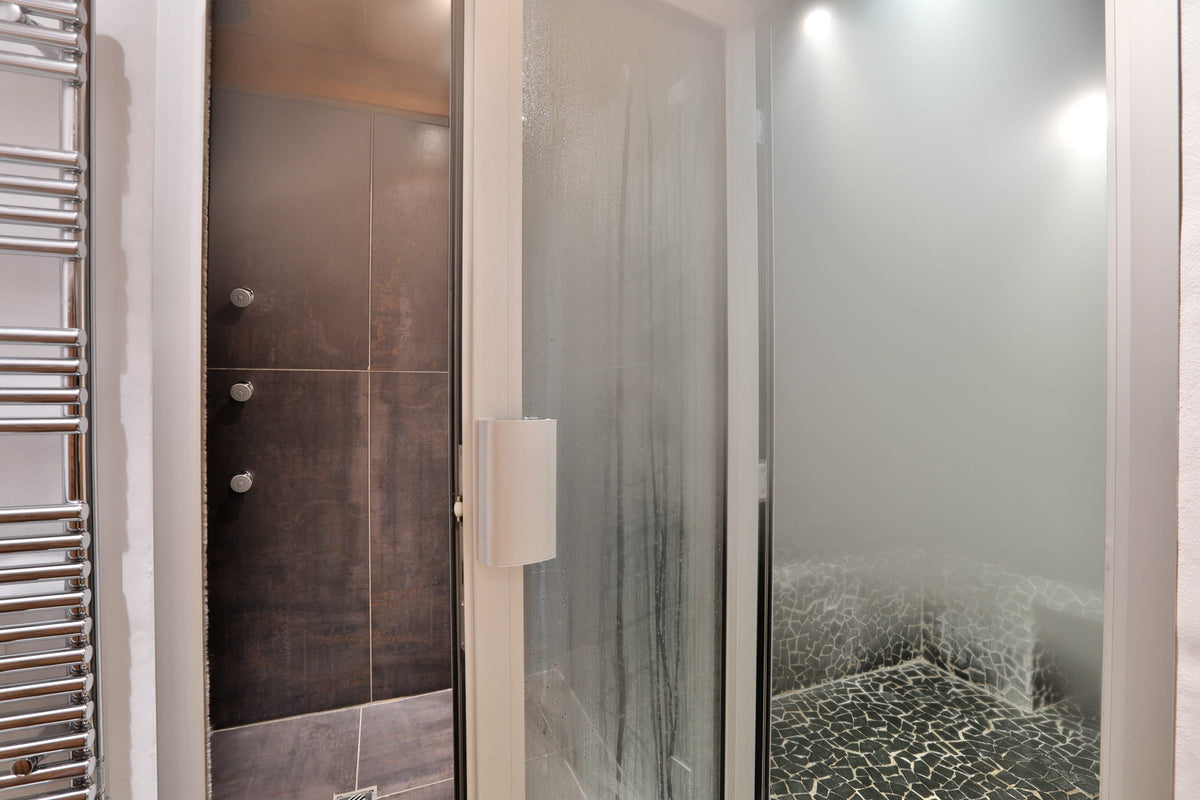 A modern indoor sauna or steam room with a frosted glass door, dark tiled interior, and bright overhead lighting.