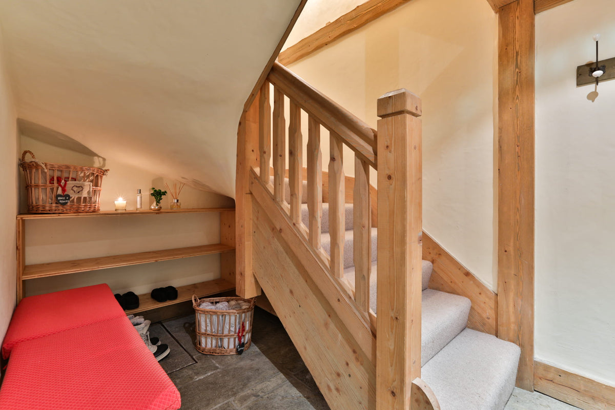 A rustic wooden staircase with light-colored wood railings and carpeted steps, set in a cozy attic or loft room with cream walls, built-in shelving, and a red cushioned bench.
