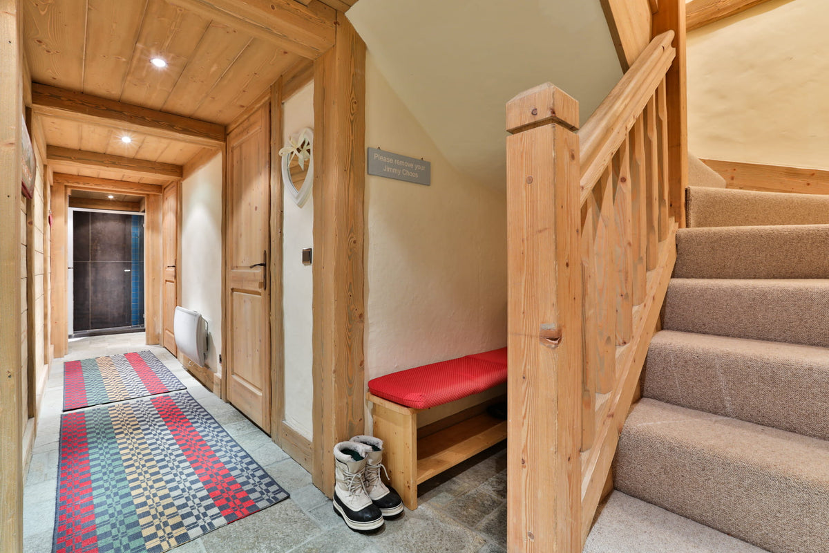 A rustic alpine-style entryway with a wooden staircase on the right, a wooden bench with a red cushion, a patterned runner rug, and a pair of winter boots on the floor.