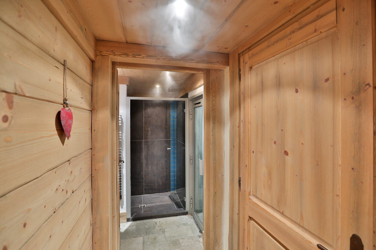 A rustic wooden interior hallway with light natural wood paneled walls and ceiling, featuring a small red heart-shaped wall hanging on the left and an open doorway revealing a modern tiled bathroom with a dark shower enclosure and glass door.