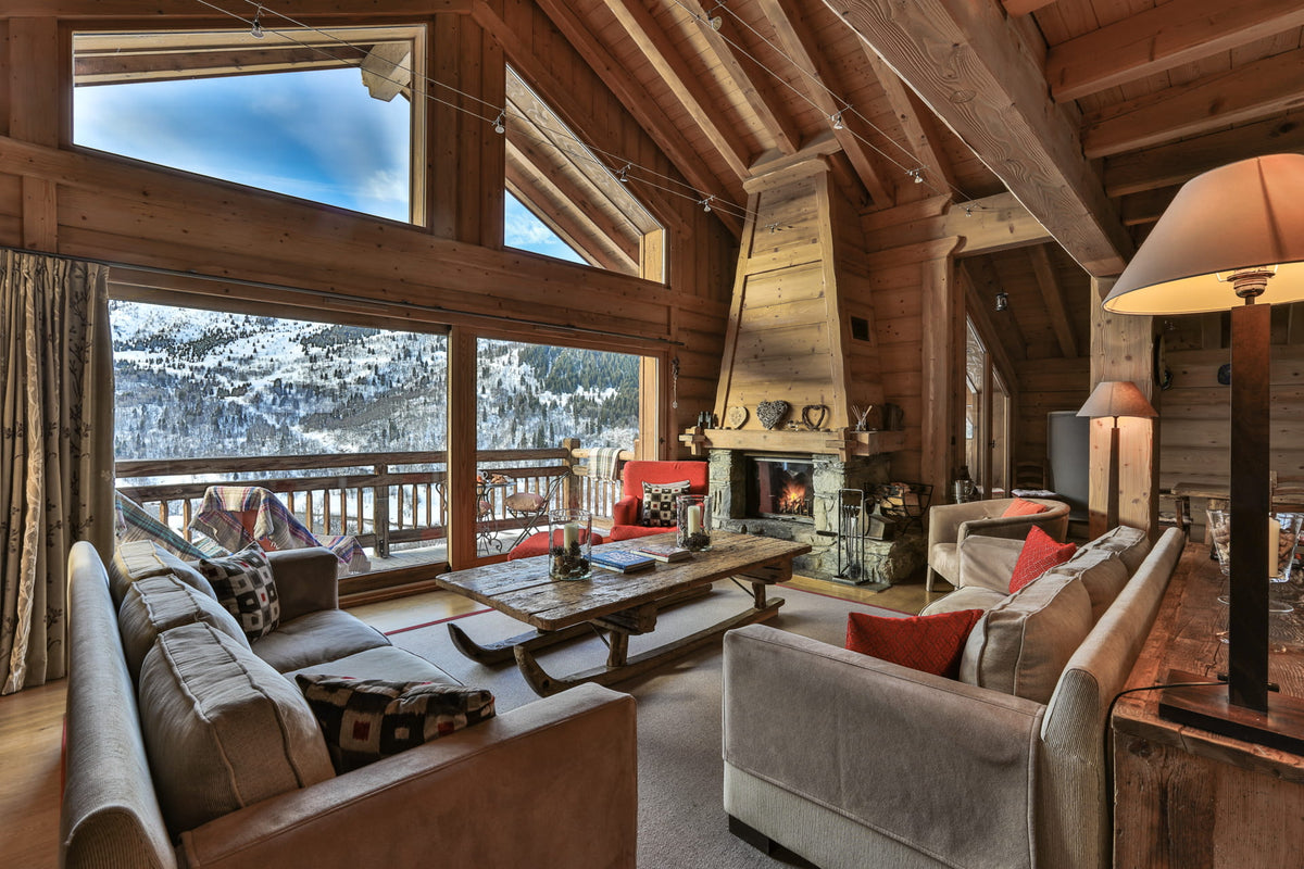 A luxurious ski chalet living room with a high wood-beamed ceiling, a large stone fireplace, and expansive windows looking out onto a snowy mountain landscape.