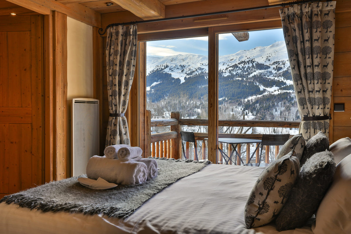 A cozy mountain-view bedroom with a neatly made bed, patterned pillows, and a large window looking out onto snowy peaks and a wooden balcony.