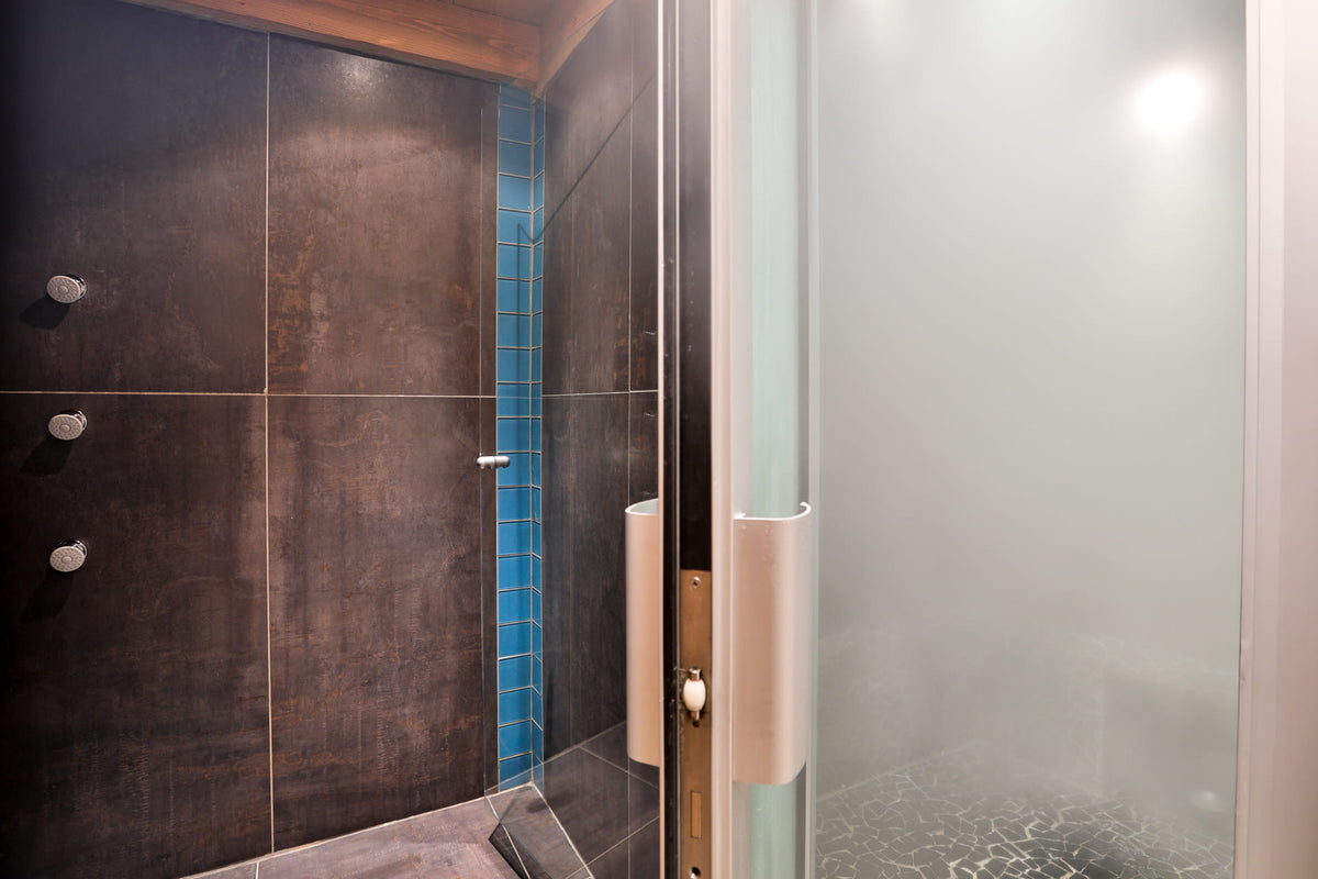 A modern walk-in shower enclosure with a frosted glass door, a beige handle, and a dark brown tiled interior featuring a vertical blue tile accent.