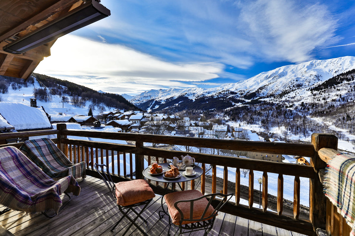 A wooden balcony overlooking a snowy mountain valley and village under a blue sky with wispy clouds. The balcony is furnished with two plaid throw blankets, a small round table with a teapot and cups, and two cushioned chairs.