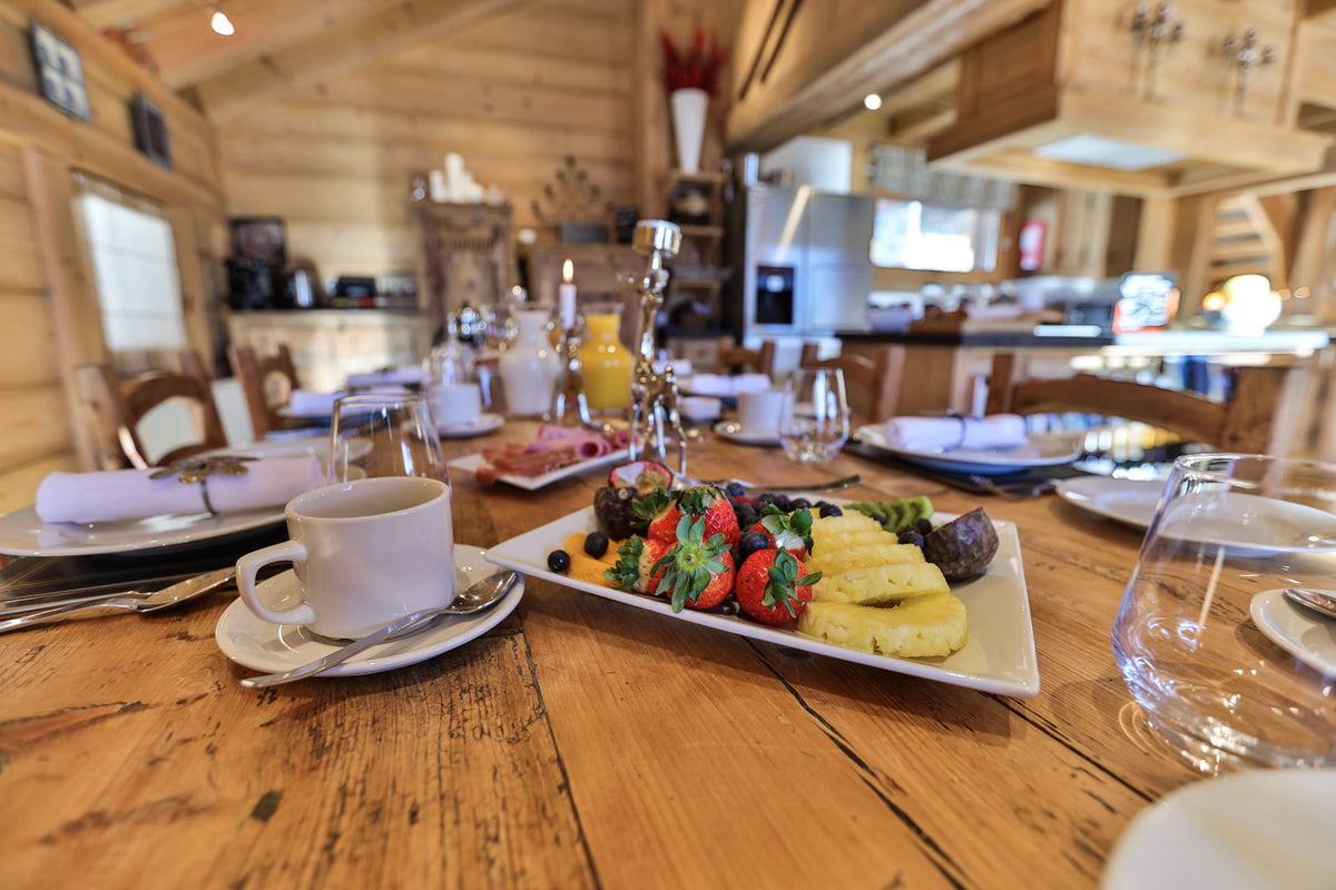 A breakfast spread on a rustic wooden table in a cabin dining area, featuring a plate with strawberries, pineapple, and sausage, alongside a white coffee cup and glasses.