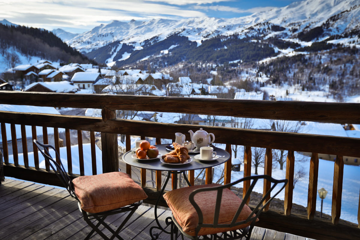A cozy outdoor balcony setup overlooking a snowy mountain village. A small round table with a teapot, cups, and pastries sits between two cushioned chairs on a wooden deck, with snow-covered peaks and forested slopes in the background.