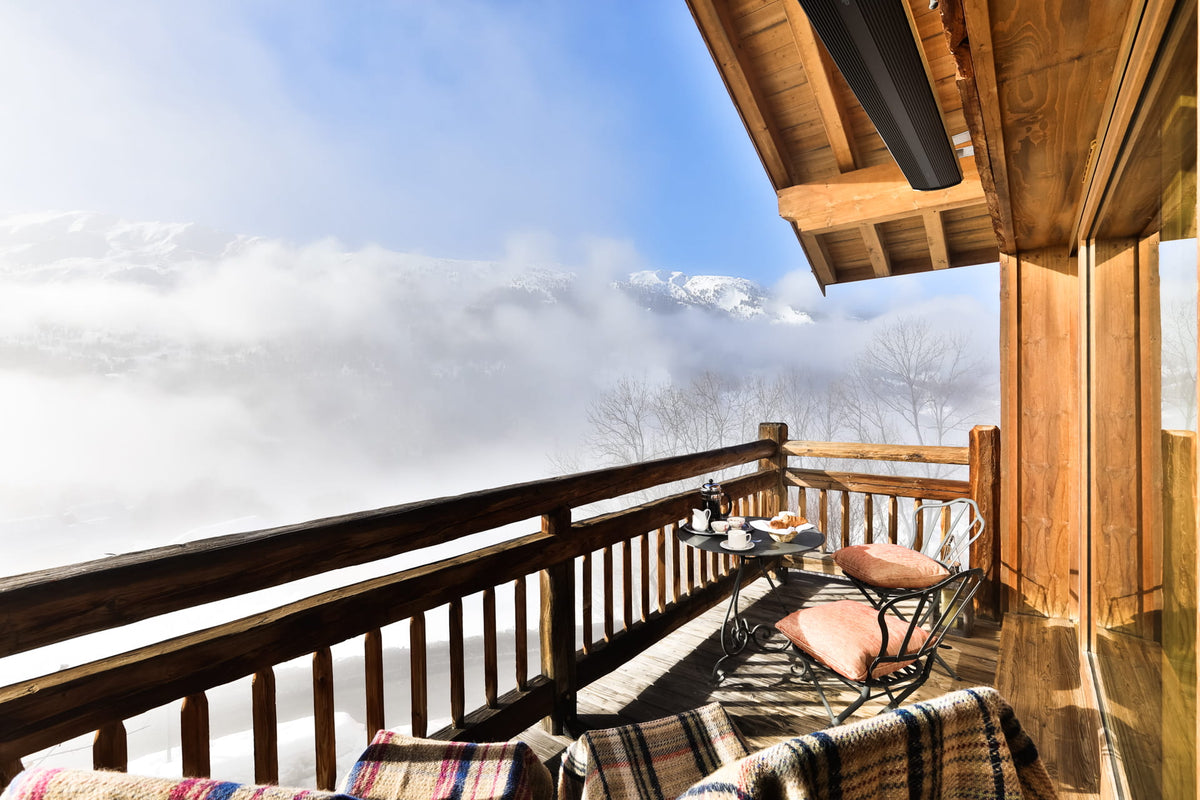 A wooden balcony with outdoor seating overlooking a misty mountain landscape under a blue sky. The balcony features warm wood paneling, a slatted railing, and cozy plaid blankets and cushions.