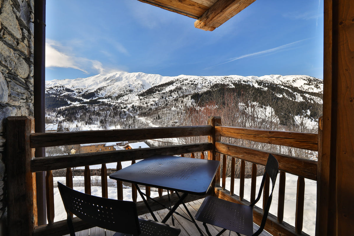 A wooden balcony with a small black table and two chairs, overlooking a snowy mountain valley under a bright blue sky.