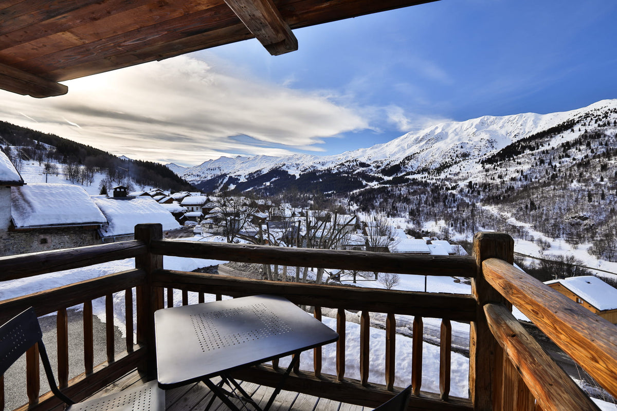 A scenic winter mountain view from a wooden balcony, with snow-covered peaks, a small village, and a blue sky in the background.