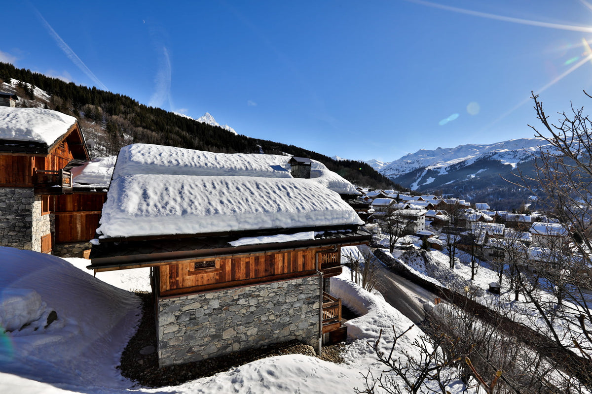 A scenic winter mountain village with snow-covered wooden chalets and distant peaks under a bright blue sky.