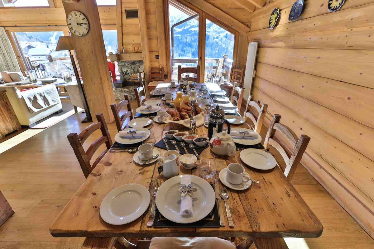 A rustic wooden dining table set for six in a cozy log cabin dining room, with white plates, cups, cutlery, and a centerpiece basket of bread and fruit, surrounded by wooden chairs.