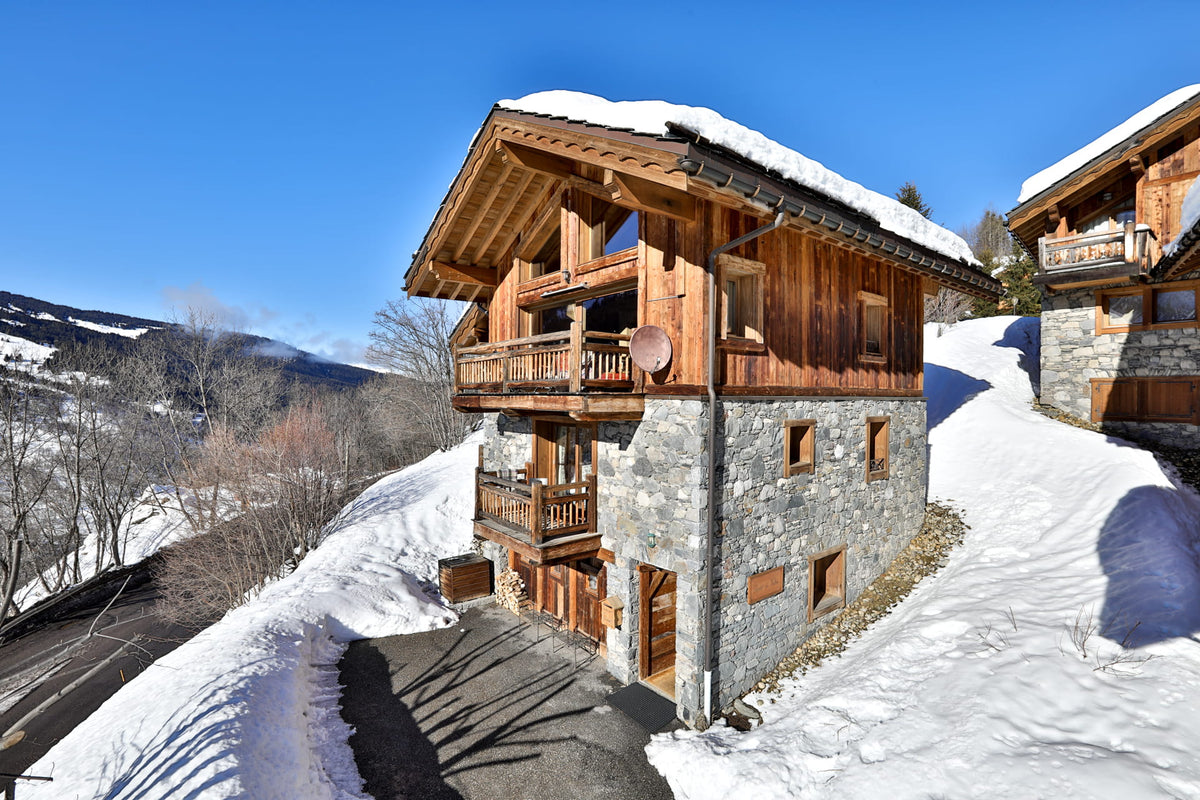 A snow-covered alpine chalet with wooden balconies and a stone lower facade, photographed on a snowy hillside under a clear blue sky.