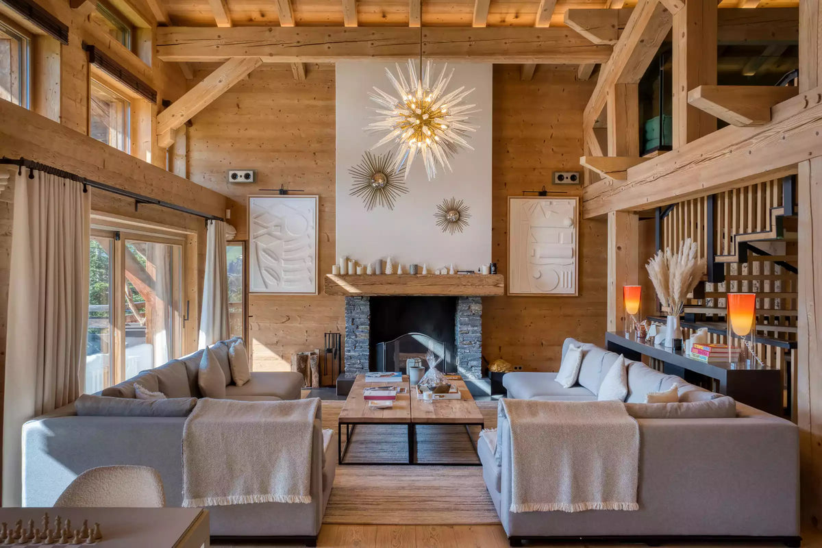 A bright, modern mountain-style living room with light gray sectional sofas, a wooden fireplace, and a large decorative sunburst chandelier hanging from exposed wooden beams.