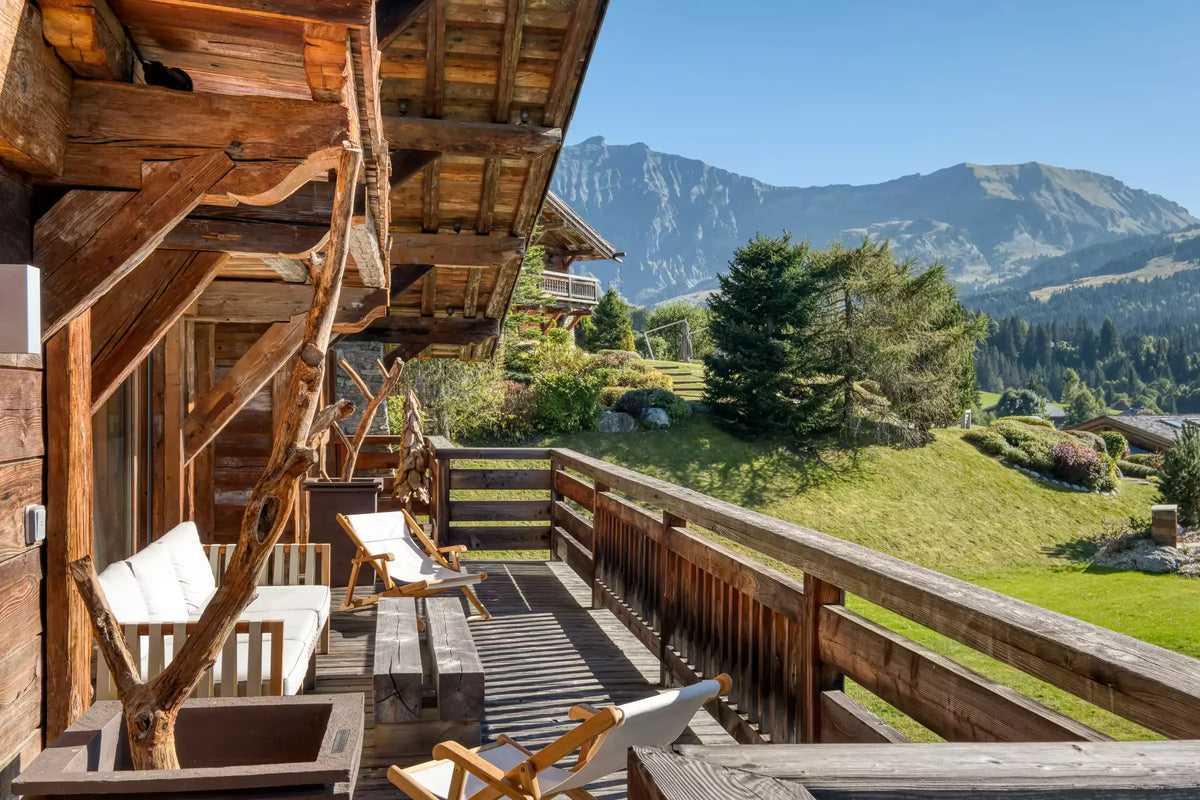 A rustic wooden chalet balcony with white cushioned chairs overlooking a lush green mountain valley and distant peaks under a clear blue sky.
