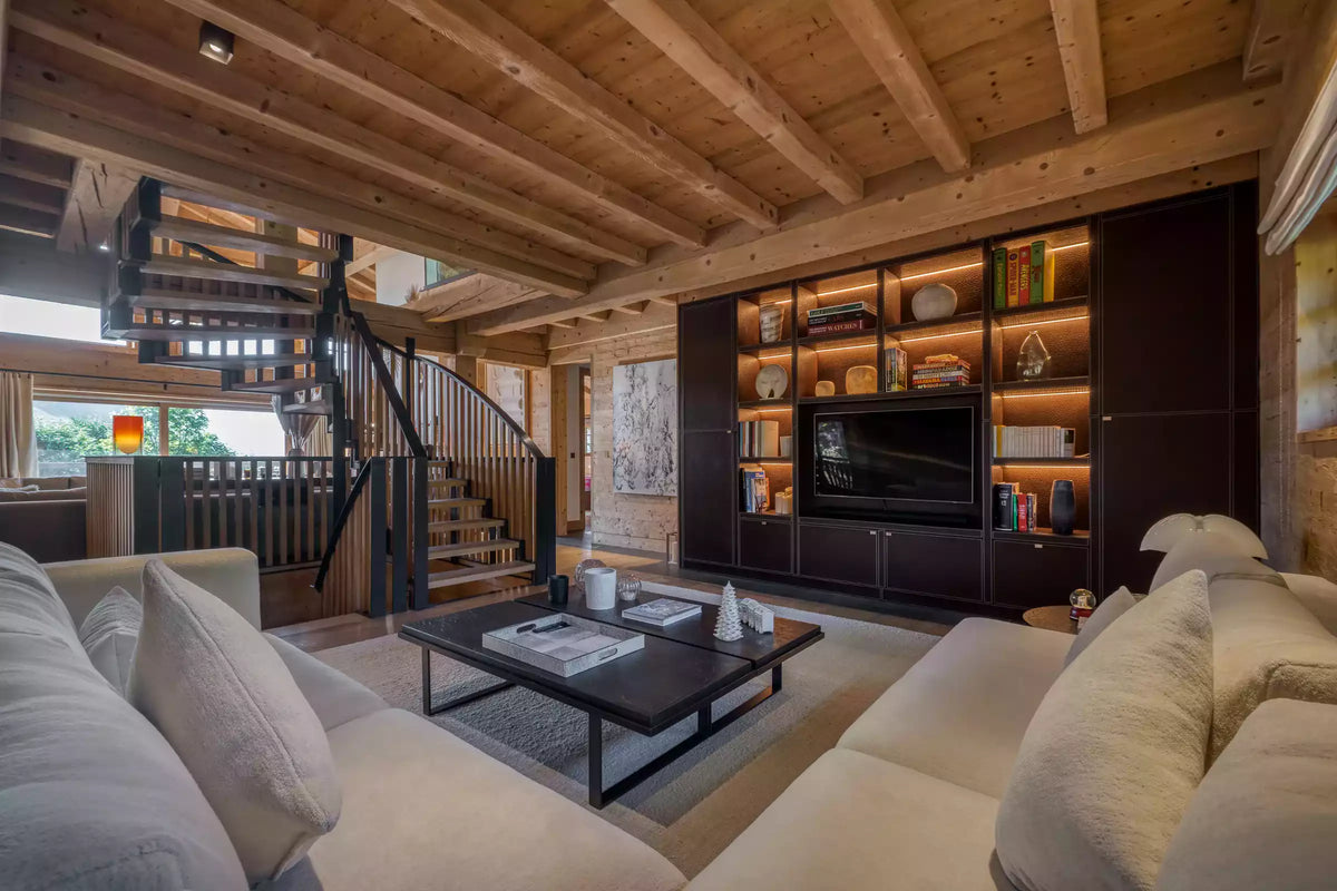 A luxury chalet living room featuring a large cream-colored sectional sofa, a dark wood coffee table, a built-in dark wood entertainment wall with a television, and a wooden staircase in the background.
