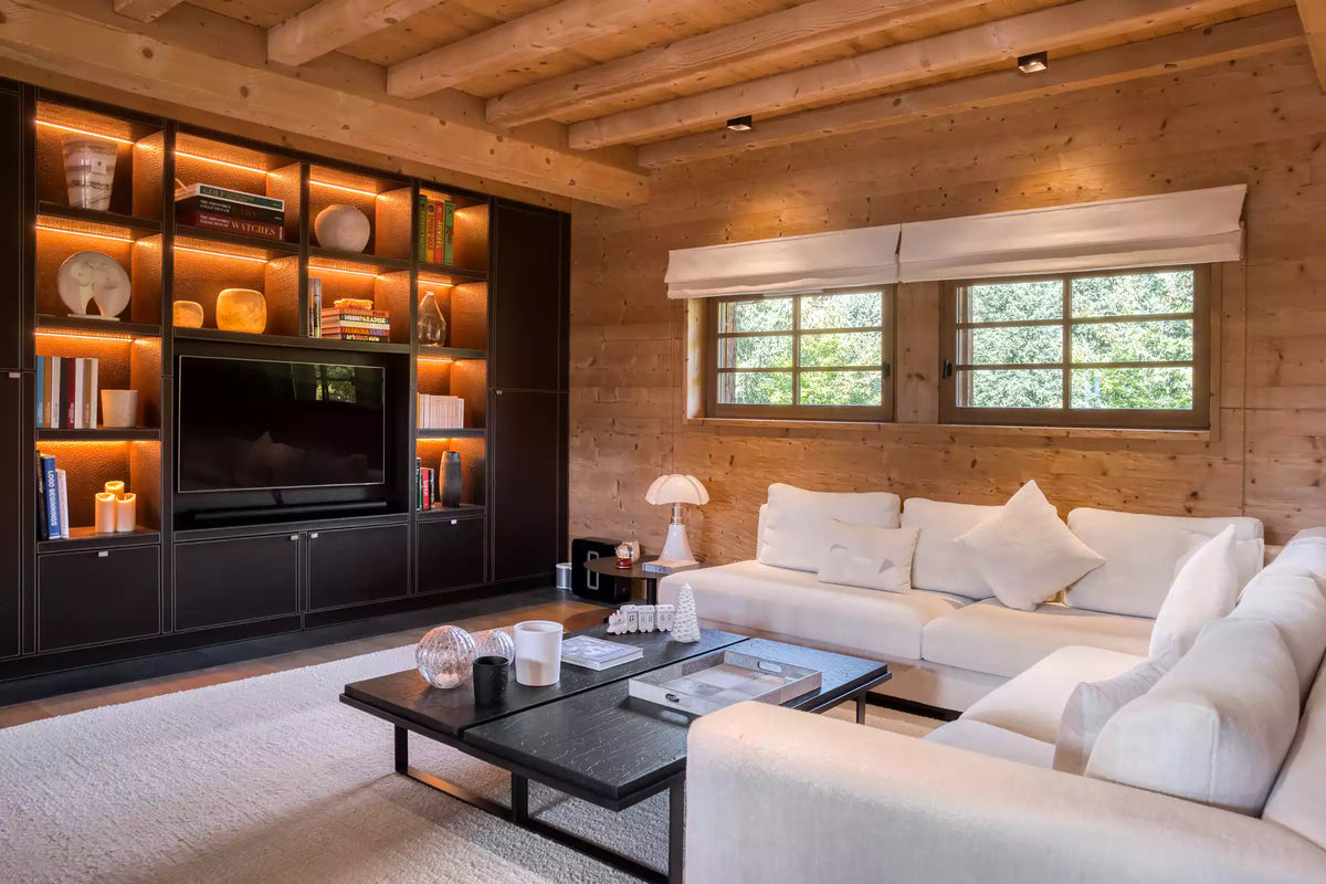 A modern living room with a white sectional sofa, a black coffee table, and a large built-in wooden media cabinet with shelves and a television. The room has warm wood-paneled walls and ceiling, two windows with white roller shades, and a light rug.