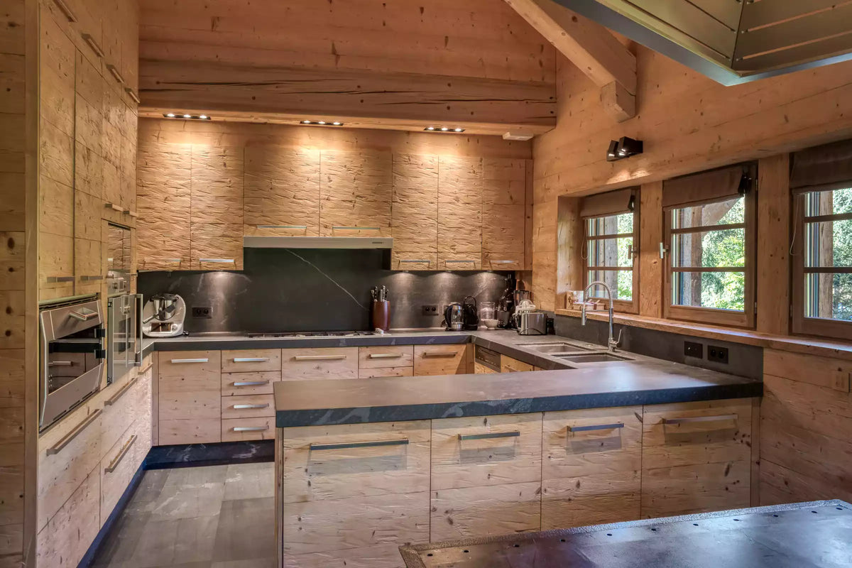 A modern kitchen interior with light wood cabinetry, dark countertops, built-in appliances, and large windows with wood frames.