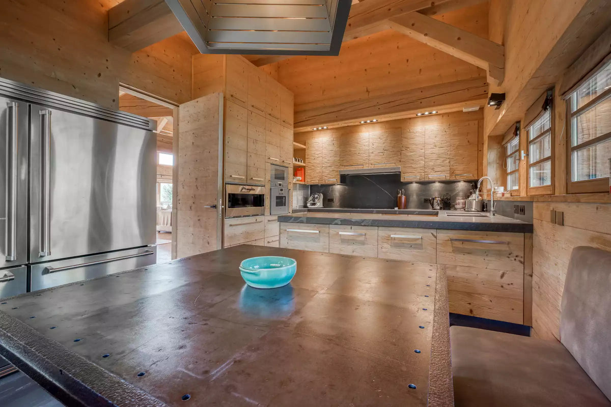 A rustic modern kitchen with light wood cabinetry, a large dark stone countertop in the foreground, stainless steel appliances, and a small translucent blue bowl on the counter.