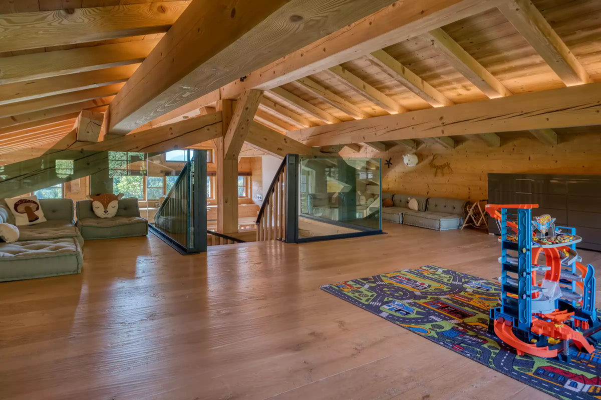 A large, open attic playroom with light wood floors and exposed wooden ceiling beams. The room features a green sectional sofa on the left, a glass railing in the center, and a colorful children's toy car race track on a patterned rug on the right.