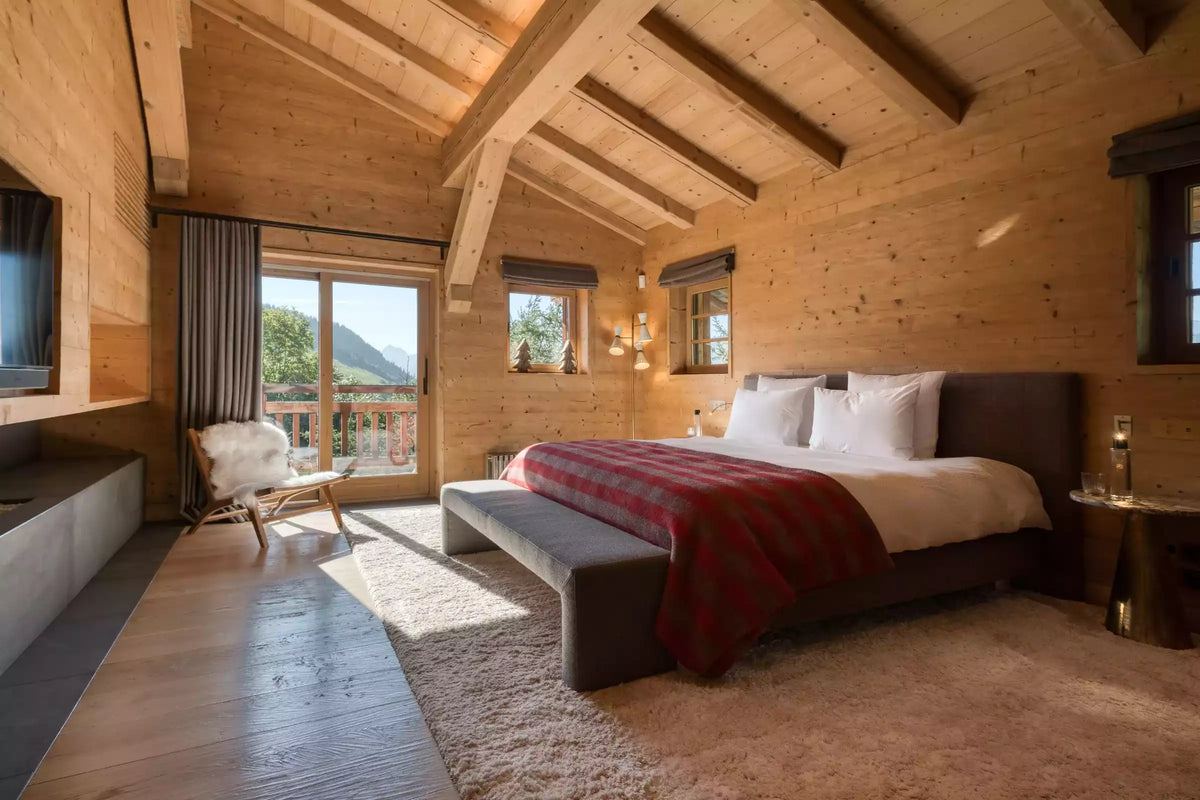 A cozy mountain chalet bedroom featuring a large bed with white linens and a red plaid throw blanket, set in a rustic wood-paneled room with a slatted wood bench, a slouchy rug, and a large window overlooking a scenic mountain view.