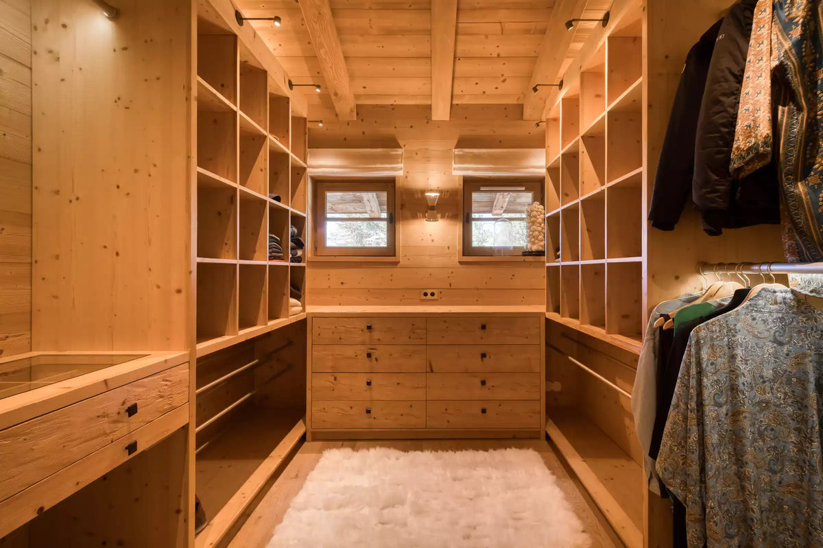 A warm wood-paneled walk-in closet with built-in shelving, a central drawer unit, two small windows, and clothing hanging on the right side.