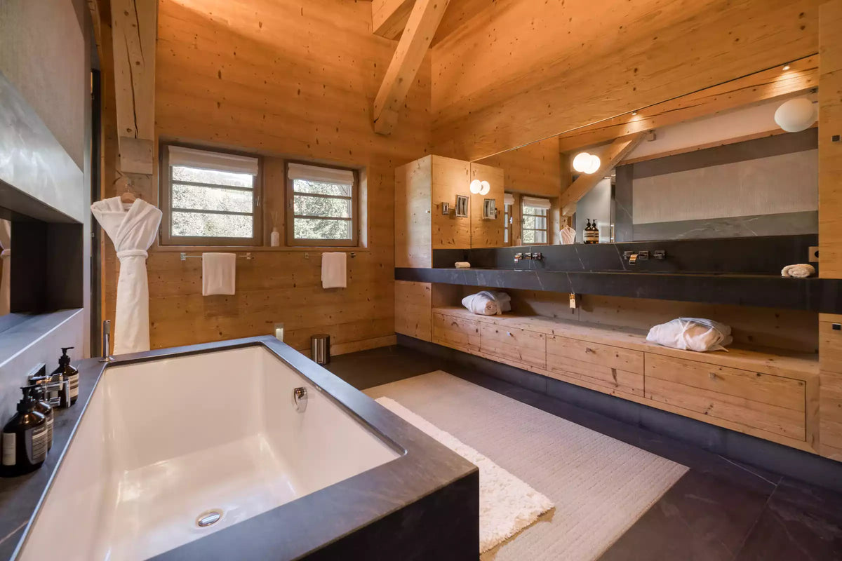 Luxury spa bathroom with a large white freestanding bathtub in the foreground, a long double vanity with dark countertop, wood cabinetry, and wood-paneled walls. A white robe hangs on the left, and two windows with blinds are visible in the background.