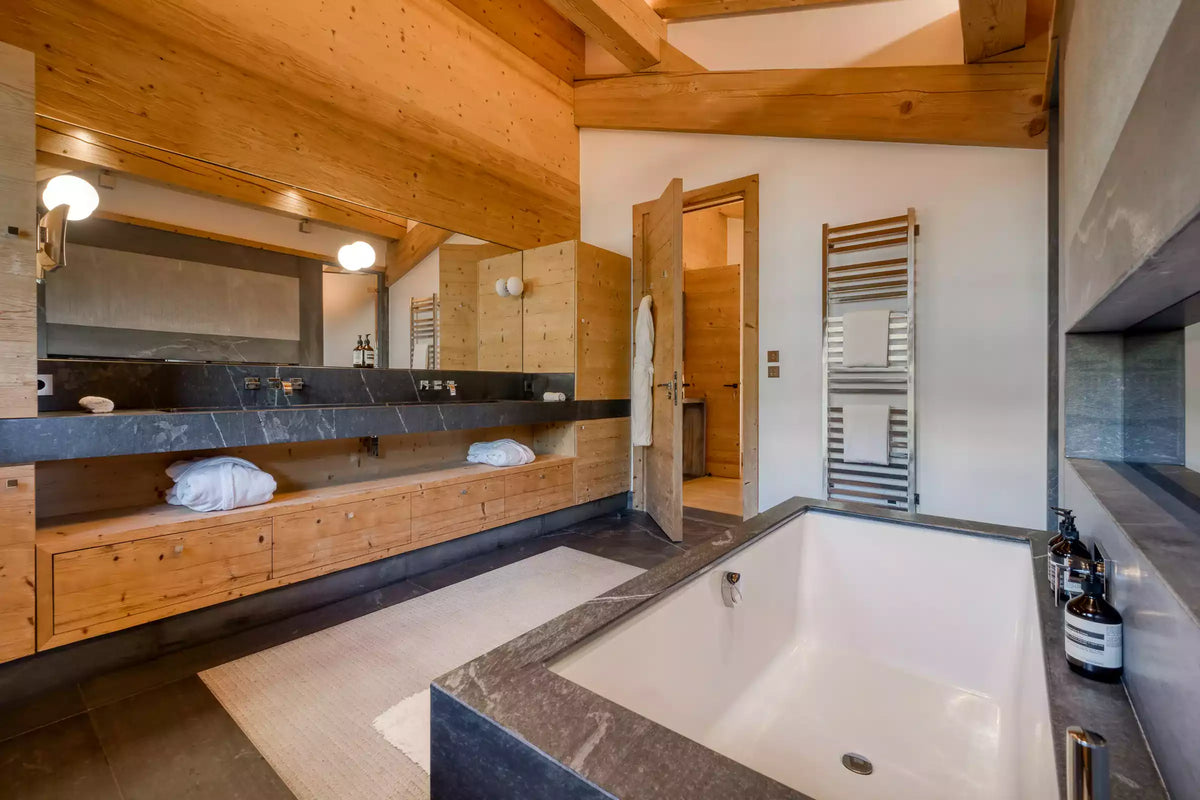Luxury bathroom interior featuring a large white freestanding bathtub with dark stone surround, a double vanity with wood cabinetry and dark countertop, wood ceiling beams, and a heated towel rail.