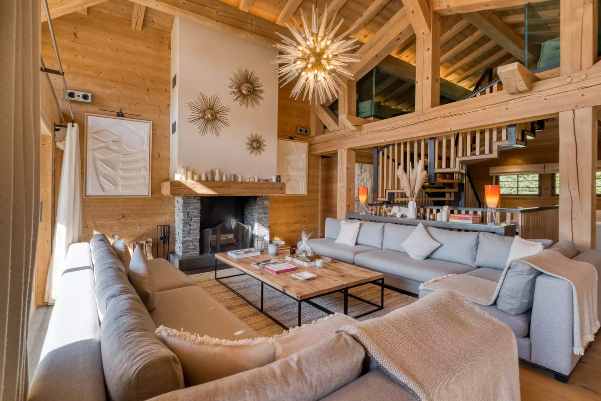 A spacious, rustic chalet-style living room with light wood walls and ceiling beams, featuring a large light gray sectional sofa, a wooden coffee table, a stone fireplace, and a starburst chandelier.