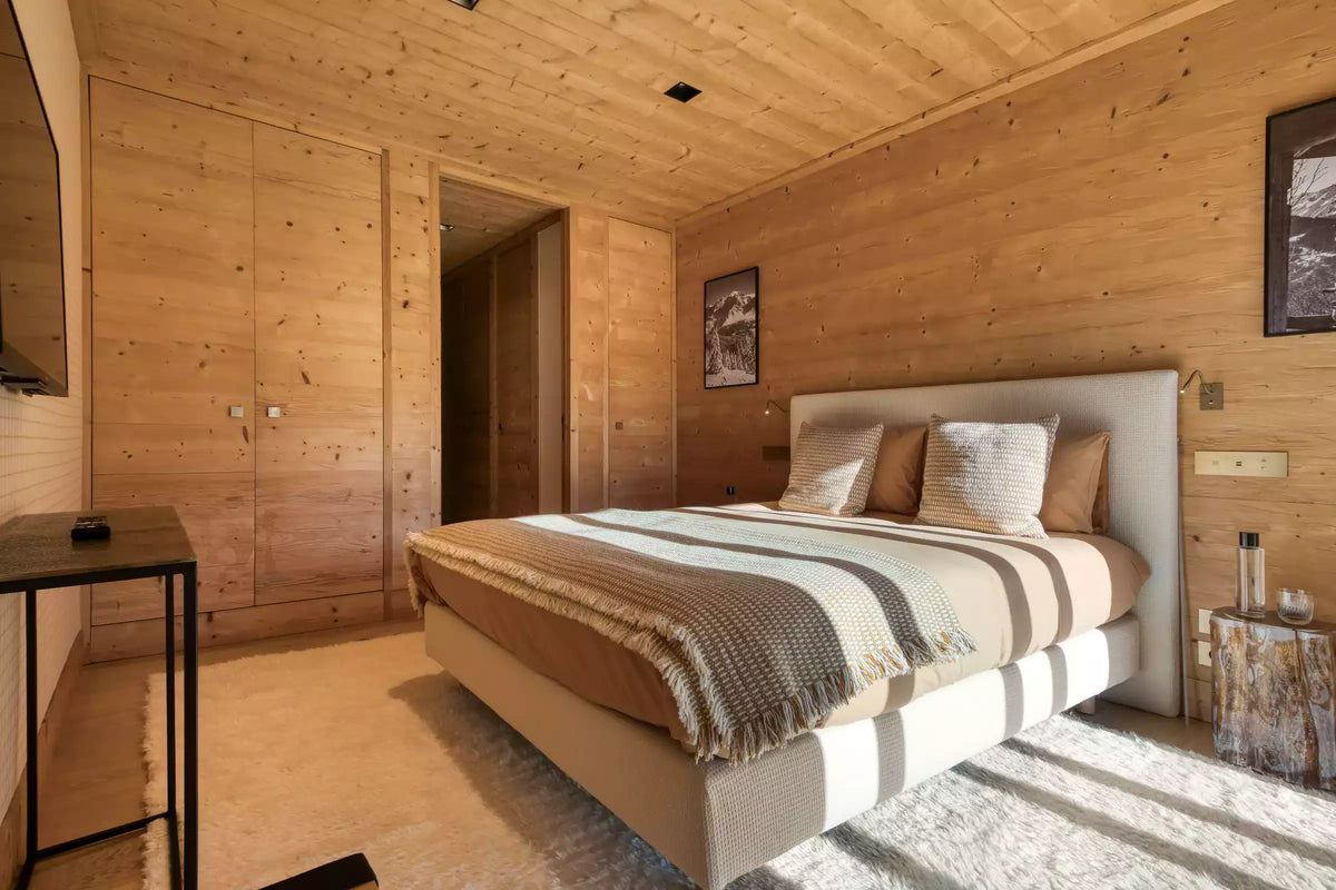A modern bedroom with light wood paneled walls and ceiling, featuring a bed with beige, brown, and white striped bedding, a white upholstered headboard, and a rustic wood nightstand.