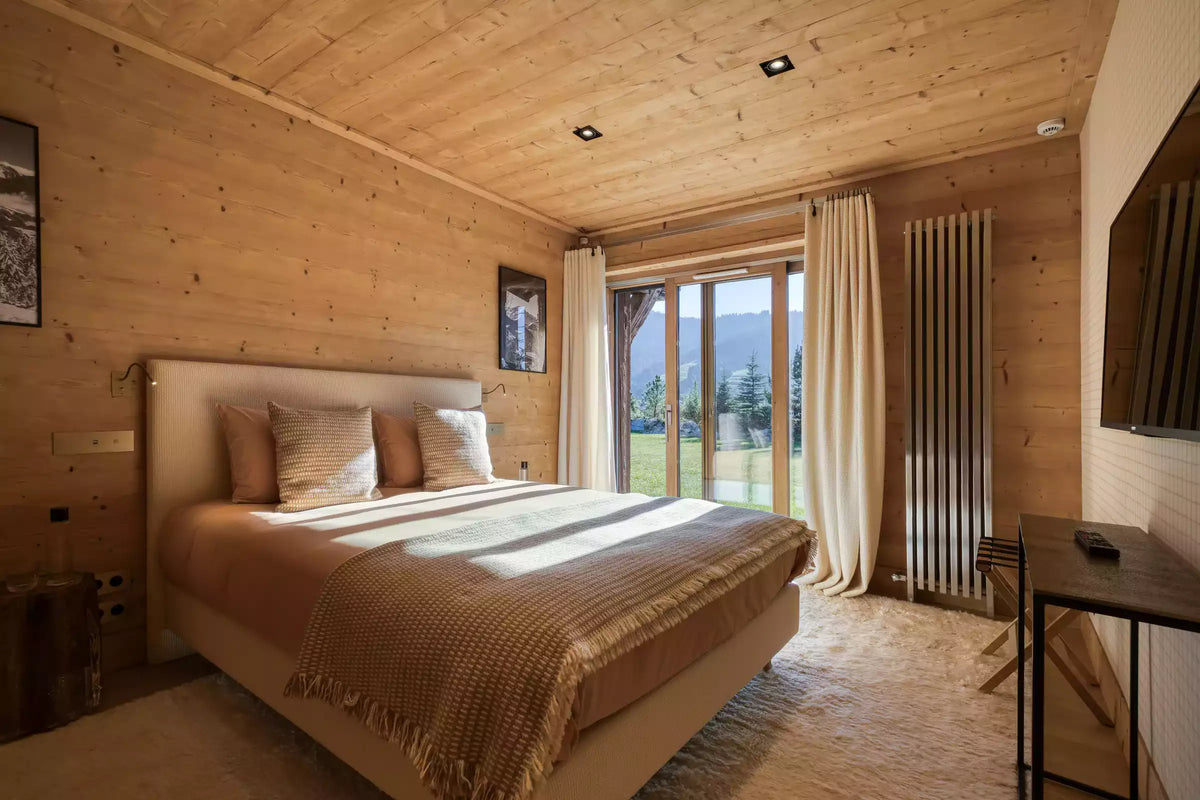 A cozy bedroom with light wood-paneled walls and ceiling, featuring a large bed dressed in beige and brown textiles, a window with cream curtains, and a small desk on the right.
