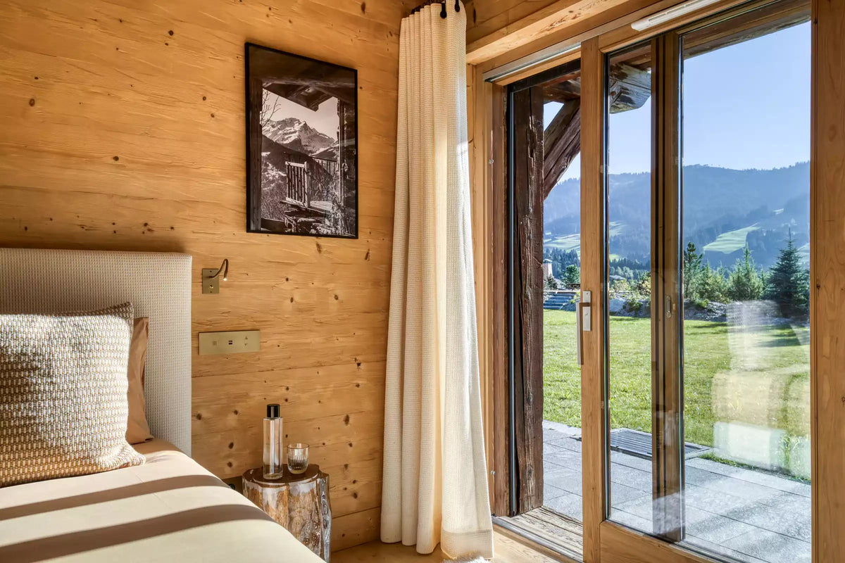 A cozy mountain chalet bedroom with light wood walls, a neatly made bed with beige and brown stripes, a small wooden side table, and a large glass sliding door looking out to a green lawn and mountain landscape.