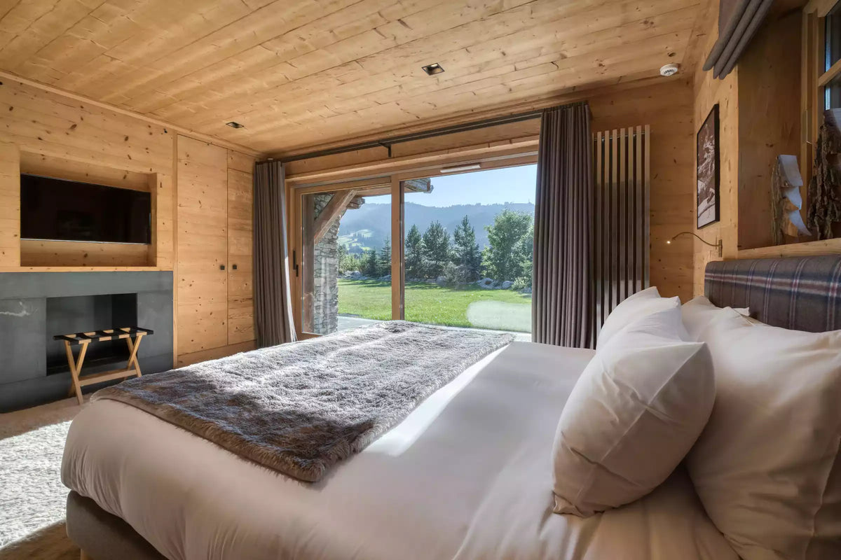 A cozy bedroom interior with a large bed dressed in white linens and a gray textured throw, set in a wood-paneled room with a sliding glass door overlooking a green outdoor landscape.