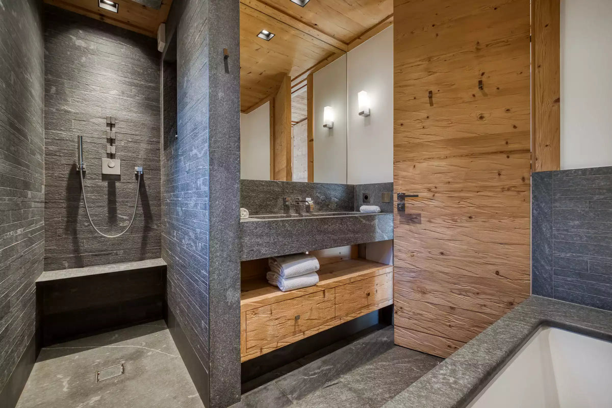 A luxury modern bathroom interior featuring a stone-tile walk-in shower, a double vanity with a dark stone countertop and open wood cabinet, a large wood-paneled door, and a white bathtub in the foreground.