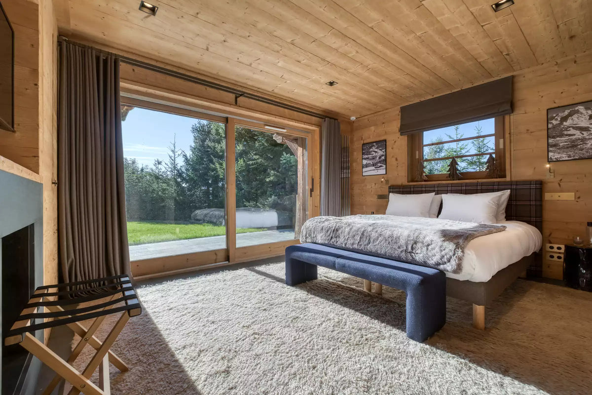 A rustic mountain chalet bedroom featuring a large bed with white bedding, a dark blue upholstered bench, wood-paneled walls and ceiling, and floor-to-ceiling sliding glass doors looking out onto a green lawn.