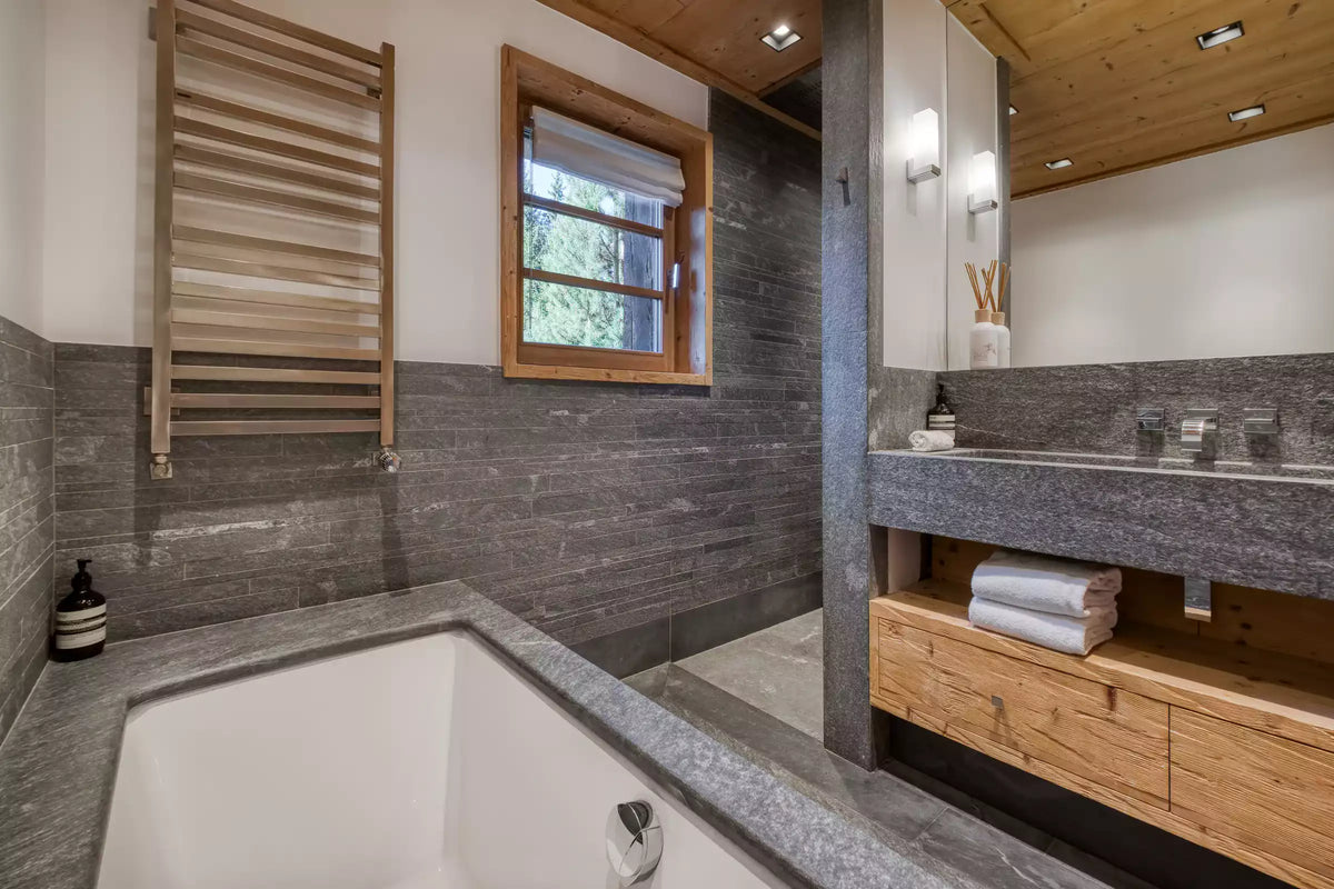 A modern bathroom interior with a white bathtub set into a gray stone wall, a wooden towel rack, a small wood-framed window, and a vanity with a gray stone countertop and wood cabinet.