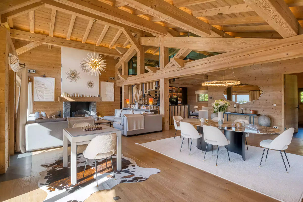 A bright, modern chalet-style dining and living room with exposed wood beams, light wood walls, a white dining table and chairs, a fireplace, and a large white rug.