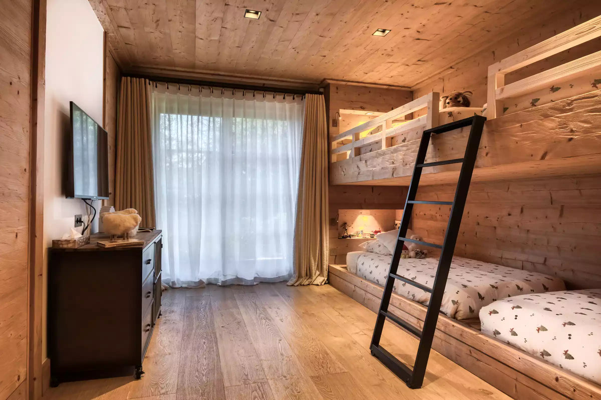 A cozy mountain-style bedroom with light wood walls and floor, featuring a built-in wooden bunk bed with a black ladder, sheer white curtains, and a dark wood dresser with a mounted TV.