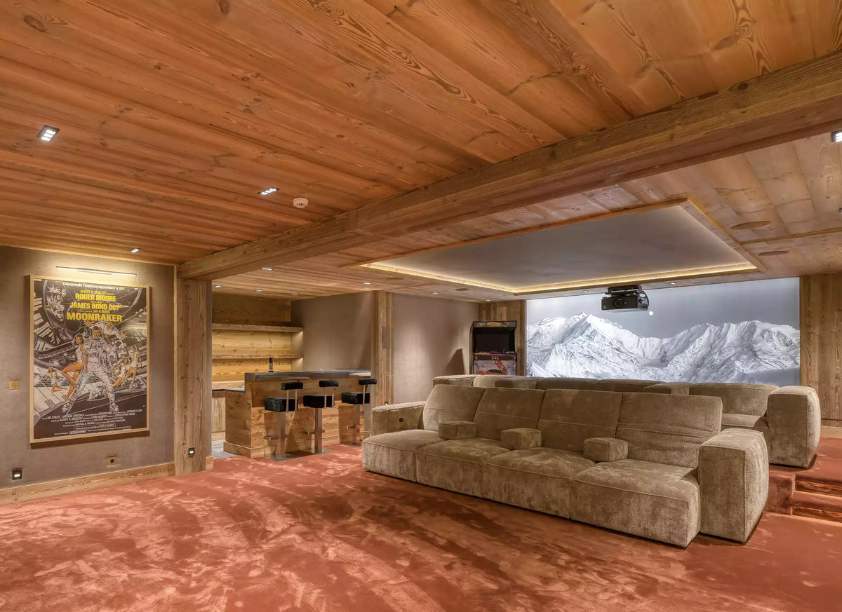 A large modern living room with a beige sectional sofa, a wood-paneled ceiling, and a large wall mural showing snowy mountains. A framed poster hangs on the left wall.