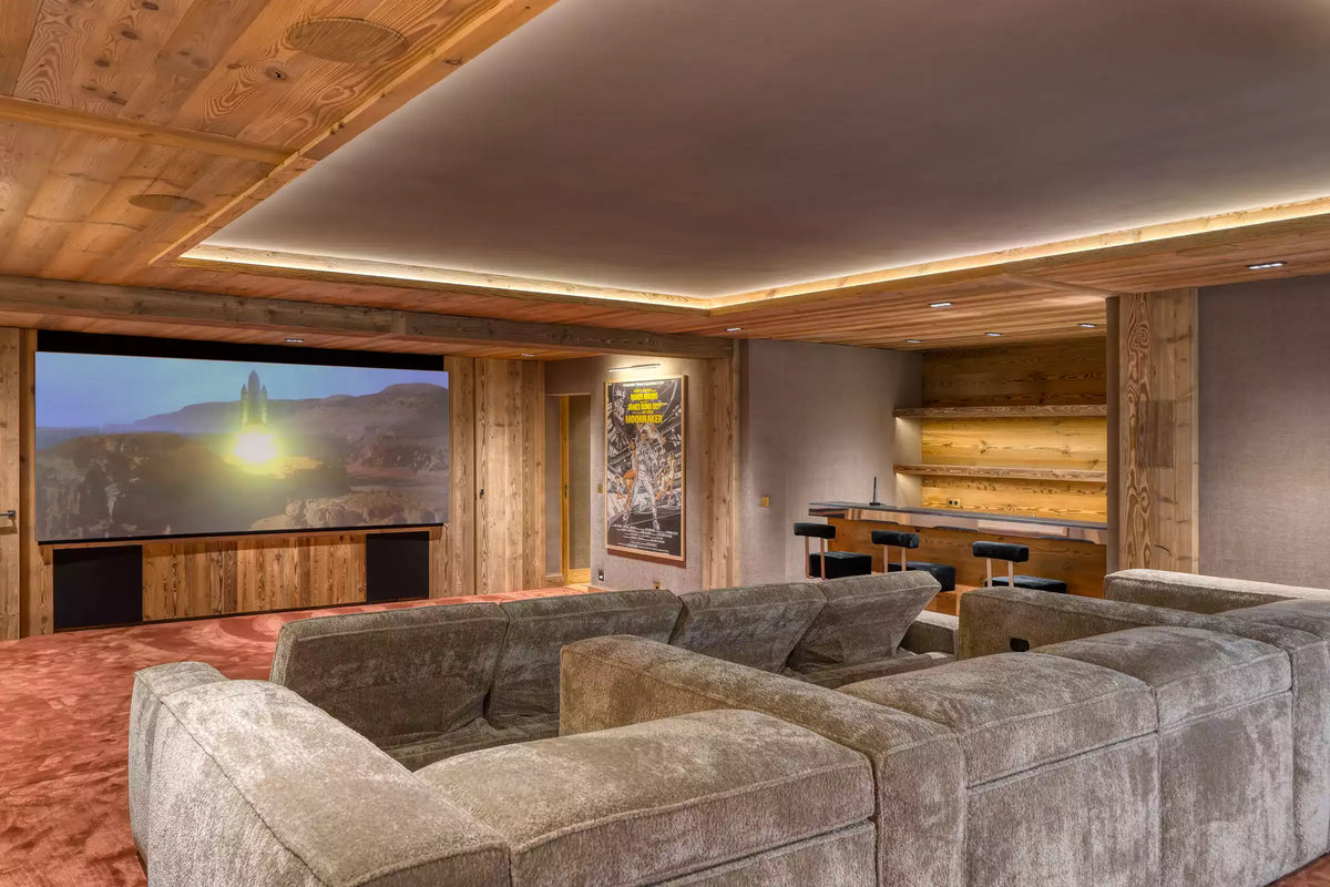 A luxury home theater room with a large U-shaped plush gray sectional sofa in the foreground, a large screen displaying a landscape, wood-paneled walls and ceiling with integrated LED lighting, and a bar area in the background.
