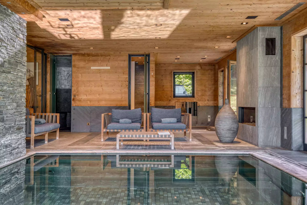 A luxury indoor poolside lounge area with two wooden armchairs, a matching coffee table, and a large decorative vase, set against a warm wood-paneled wall and stone accents.