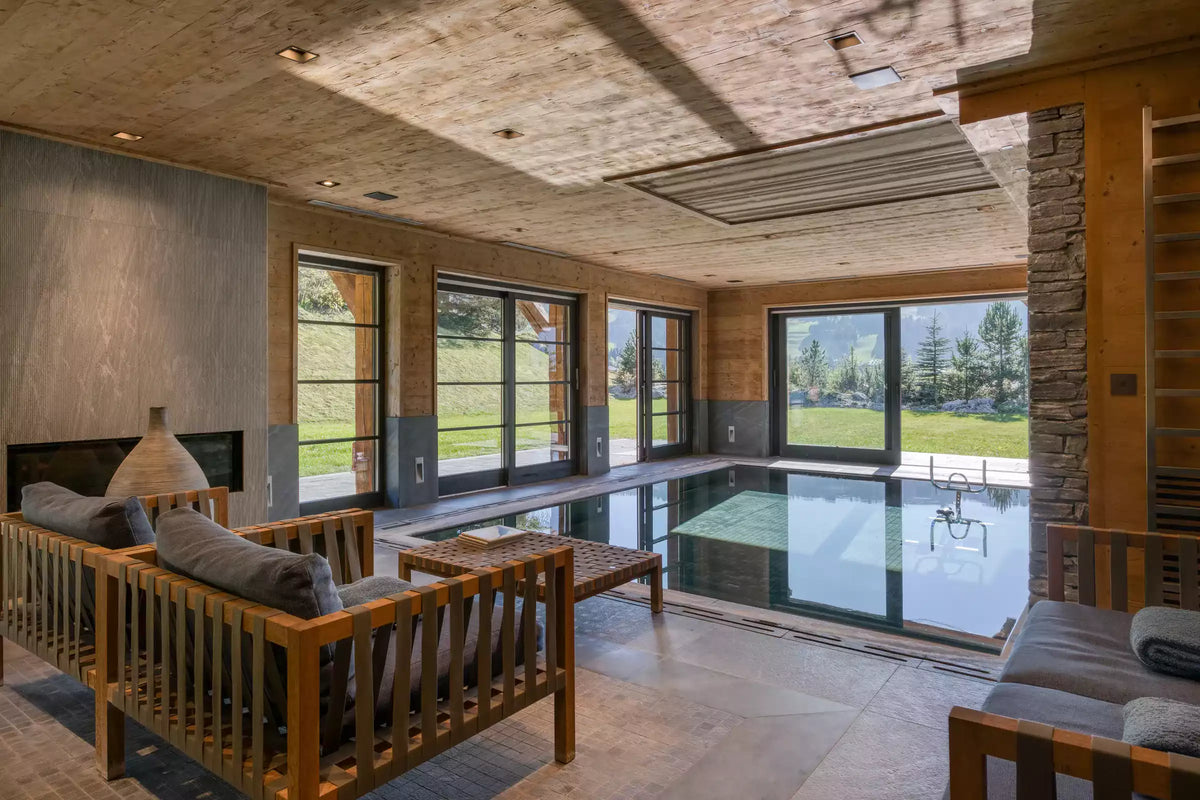 A luxury modern indoor poolside lounge area with wooden lounge chairs, a low wooden coffee table, and large floor-to-ceiling windows overlooking a green outdoor lawn, set against a rustic wood and stone interior.