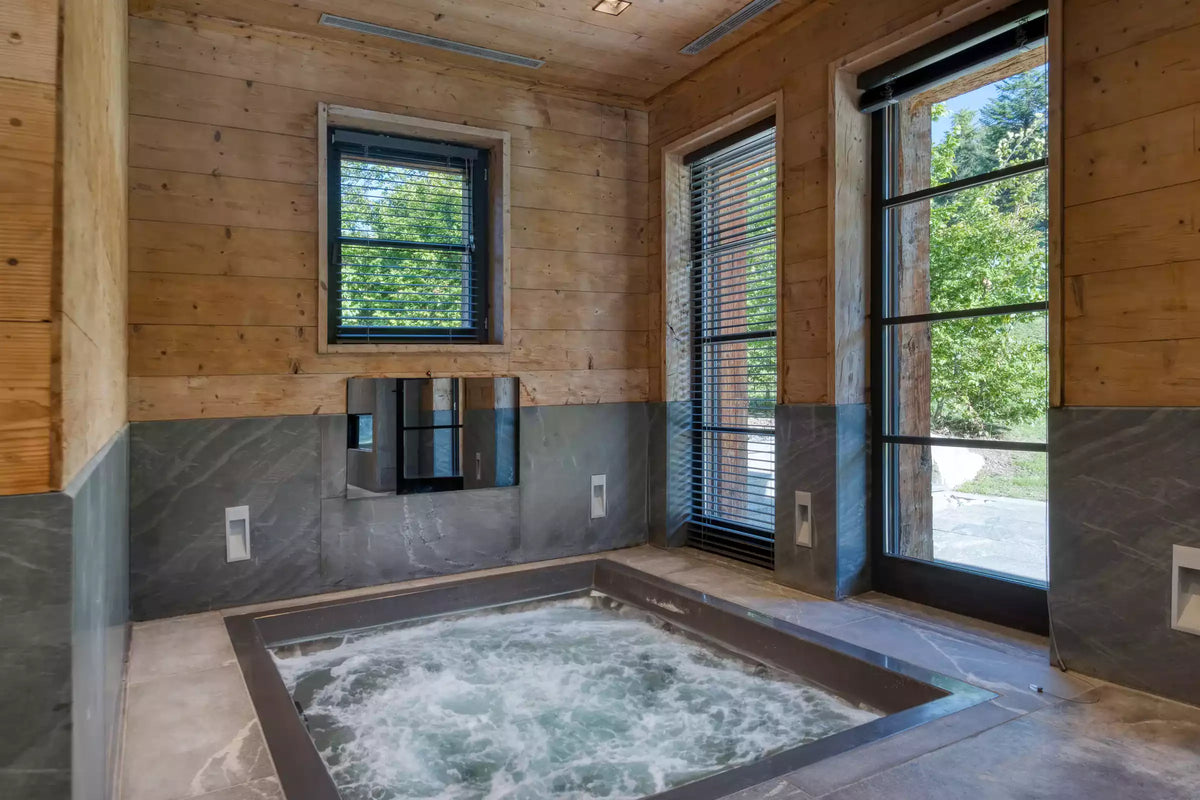 An indoor hot spring or bubbling hot tub set into a rustic wooden cabin room with wood-paneled walls, gray stone lower walls, and large windows looking out to green trees.