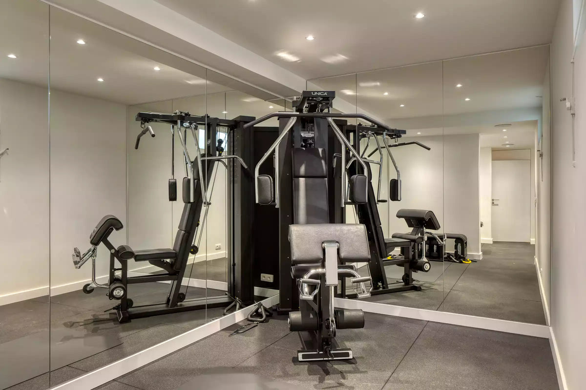 A black and gray multi-functional home gym cable machine with a front attachment pad, set in a clean, modern fitness room with mirrored walls and gray flooring.