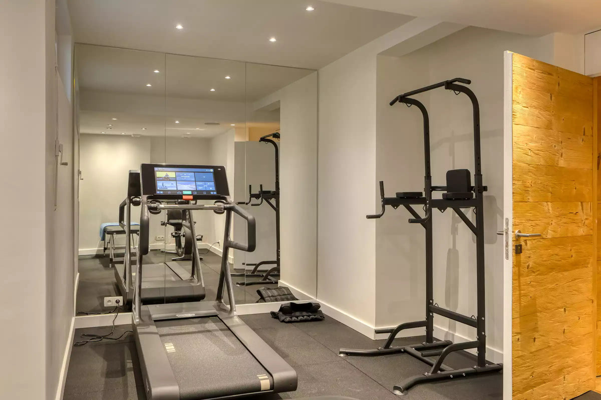 A home gym interior featuring a treadmill with a digital display, a dual pull-up bar strength training stand, and exercise equipment reflected in a large wall mirror. The room has light gray walls, dark gray flooring, and a light wood door on the right.
