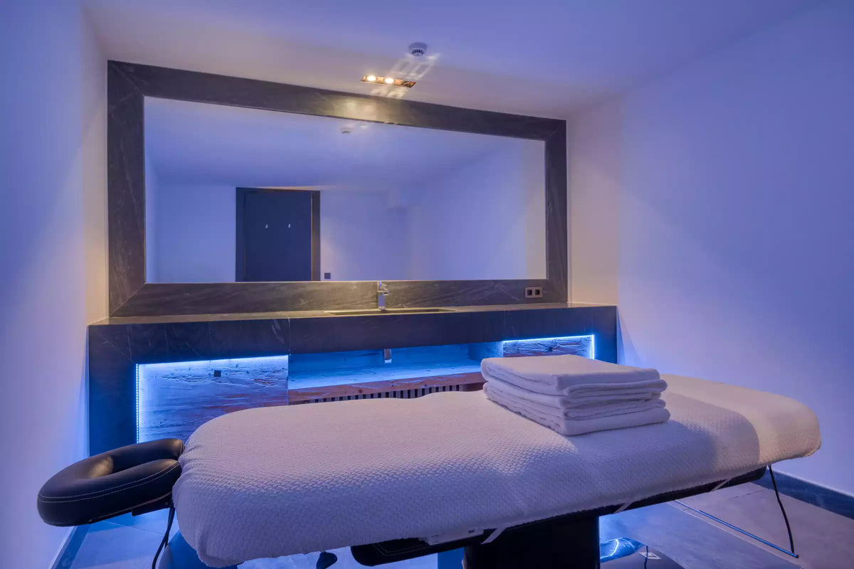 A spa treatment table with white linens and folded towels, positioned in front of a large wall mirror in a blue-lit treatment room.